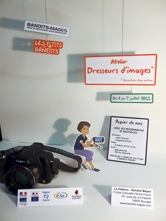 Photo utilisée pour l&rsquo;affiche et le flyer. Les différents éléments (date, logo, contenus, contact, etc, sont imprimé sur du papier, découpé et agencés grâce à des fils ou posé sur la table. Un personnage imprimé sur du papier découpé est assis sur un vrai appareil photo. Un éclairage permet un jeu d&rsquo;ombre derrière les différents éléments afin de faire ressortir la profondeur.)